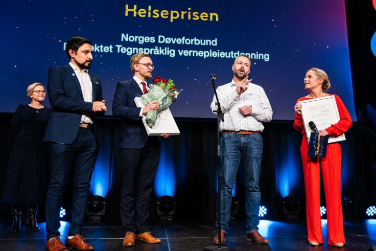 Bilde av tre menn (generalsekretær og prosjektkoordinator i Norges Døveforbund) og prosjektleder Finn Arild Thordarson) som mottar Helseprisen 2022. På scenen sammen med dem står programleder Solveig Kloppen og helseminsiter Ingvild Kjerkol.